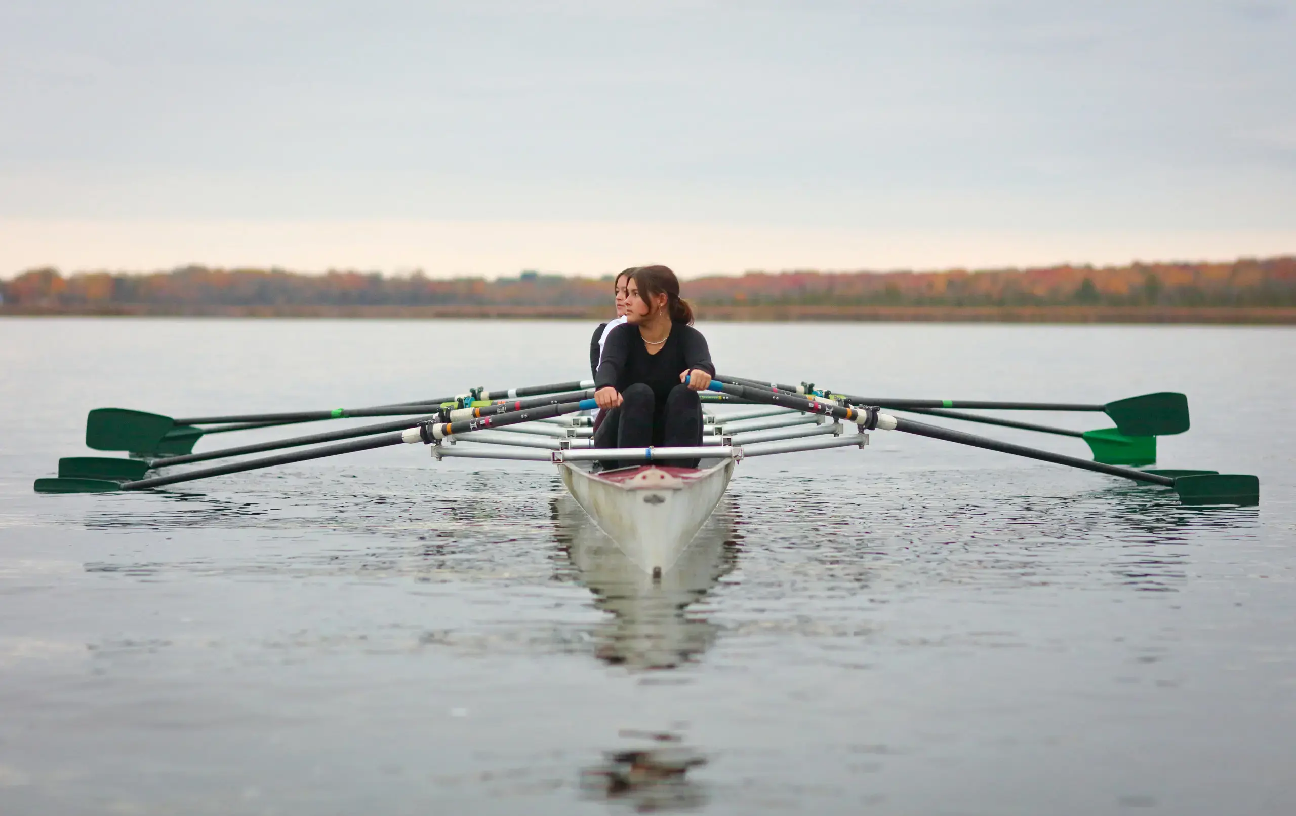 Two students in a racing shell row across a calm lake.