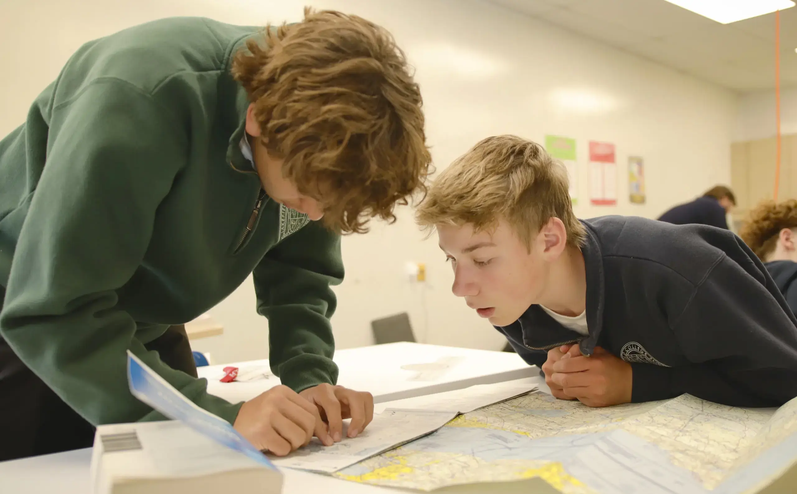 One student bending over his classmate at a desk, both working on a project