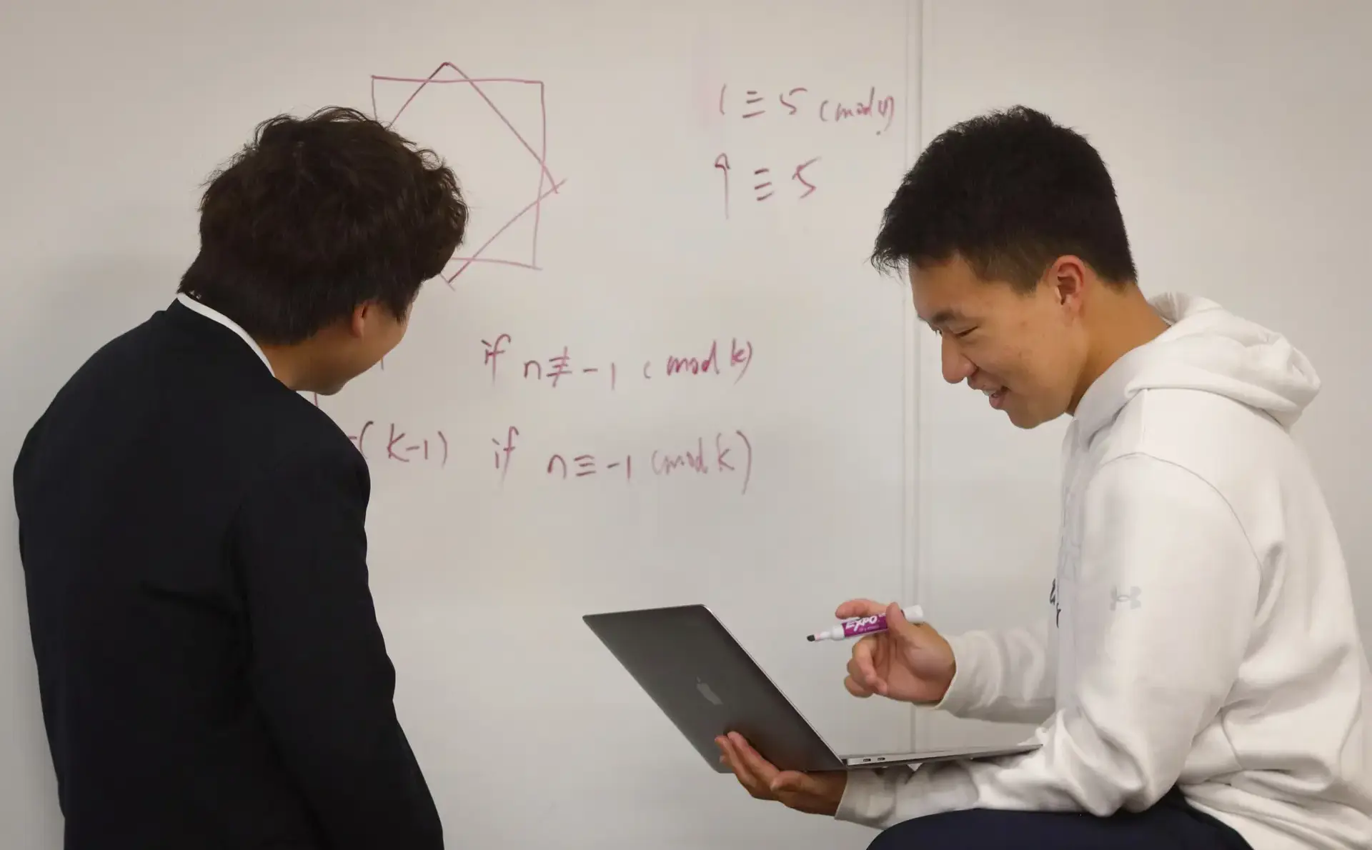 Students at whiteboard, one holding a laptop