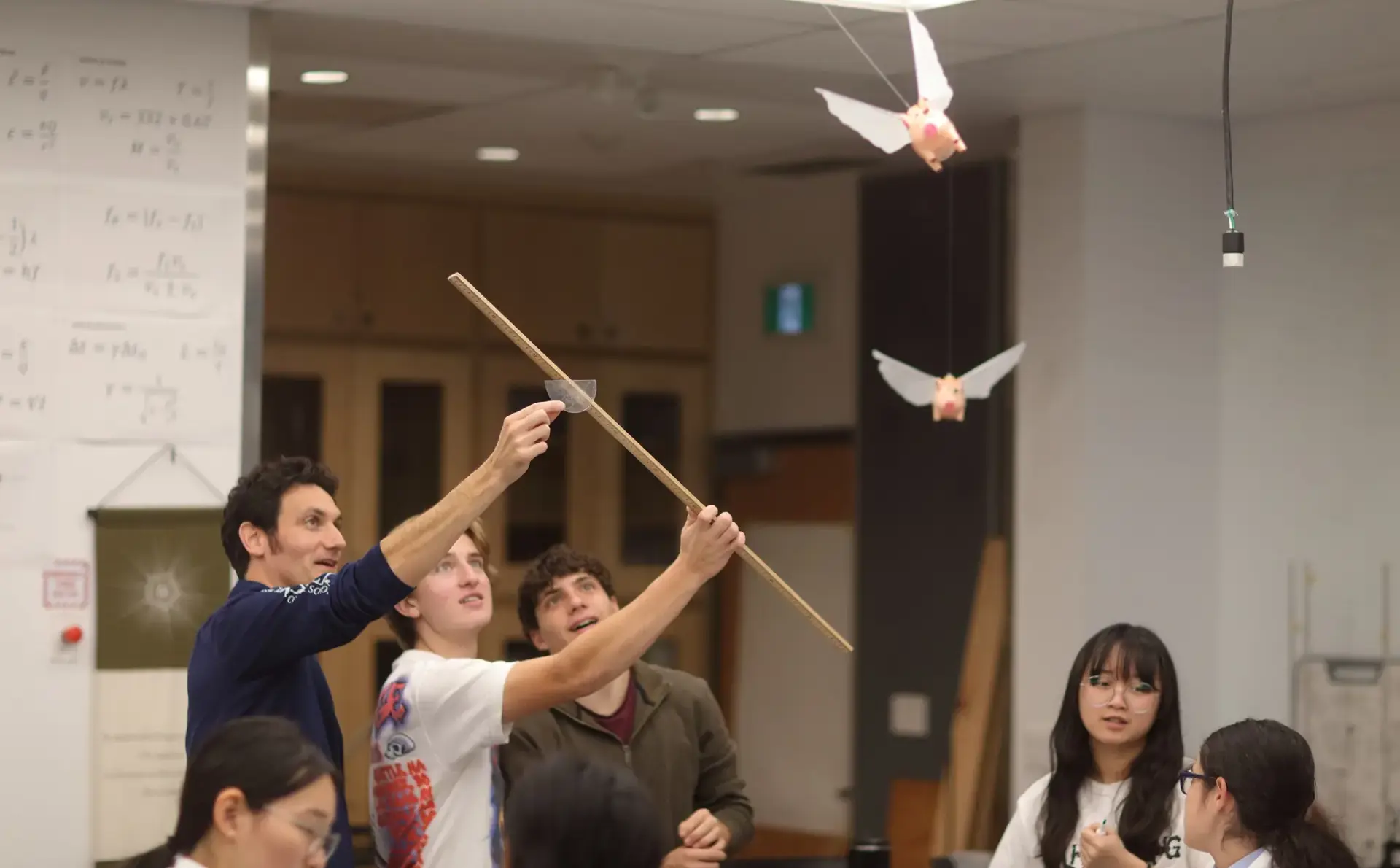A teacher and two students use a meter stick and a protractor to measure the angle and flight path of winged toy pigs hanging from the ceiling.