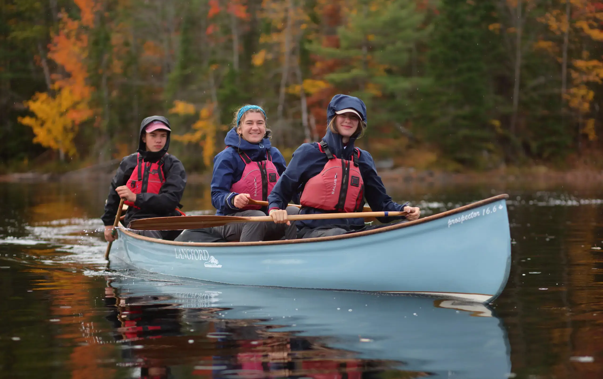 Three smiling students in life jackets and rain gear sit in a blue 'Langford' canoe on a calm lake, paddling together.