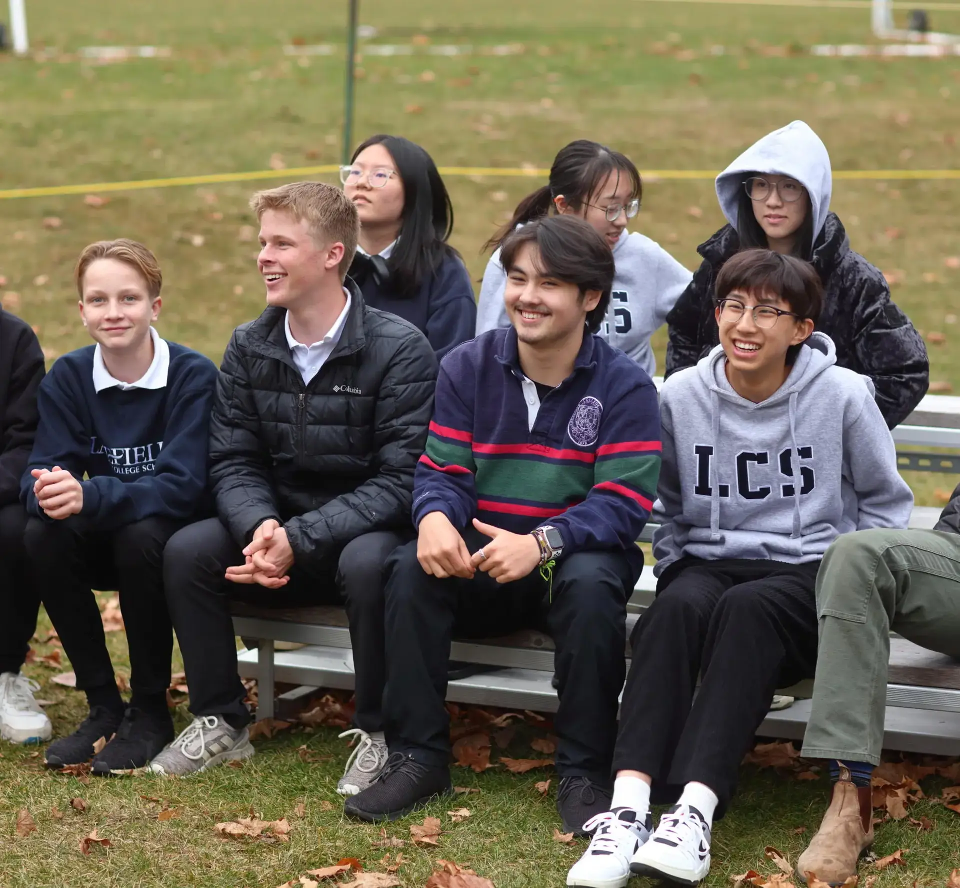 A group of students and a teacher sitting on a wooden bench outdoors on a grassy field.