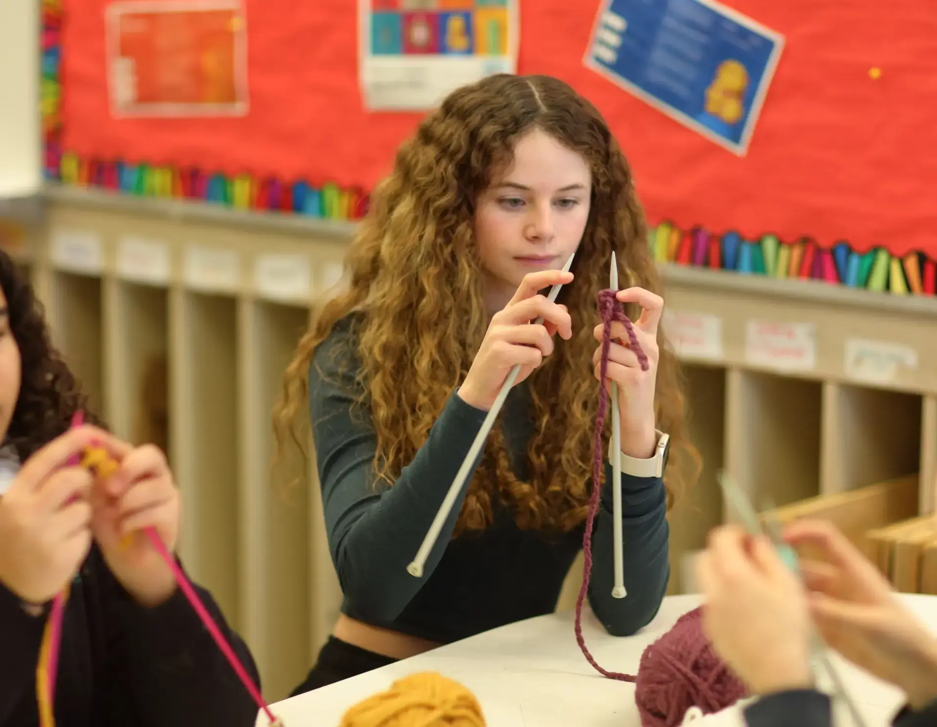 A student with long curly hair sits at a white table, focused on knitting with purple yarn and grey needles. 