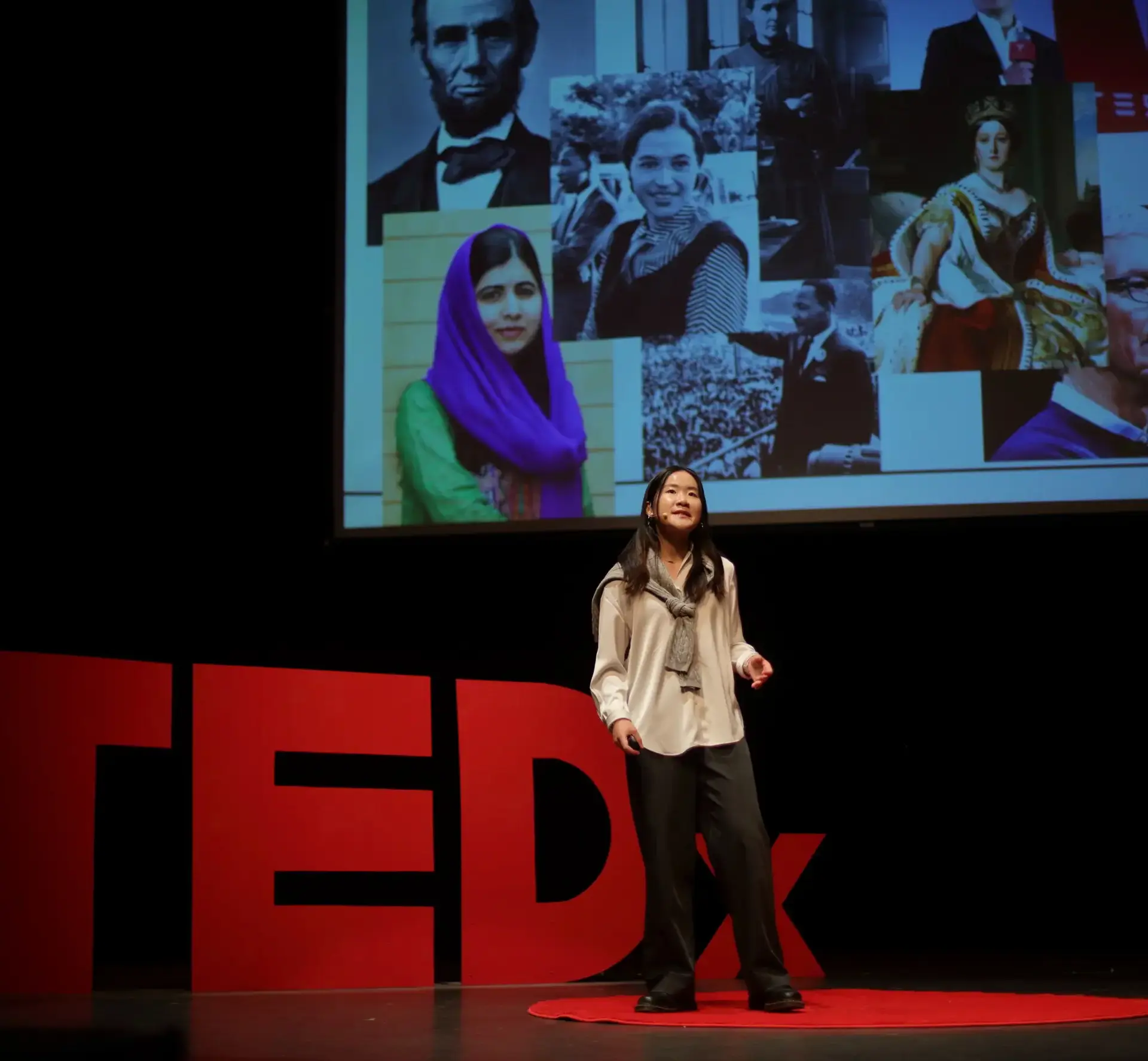 A student stands on a stage in front of a large red 'TEDx' sign and a screen displaying images of historical and influential figures, delivering a presentation.