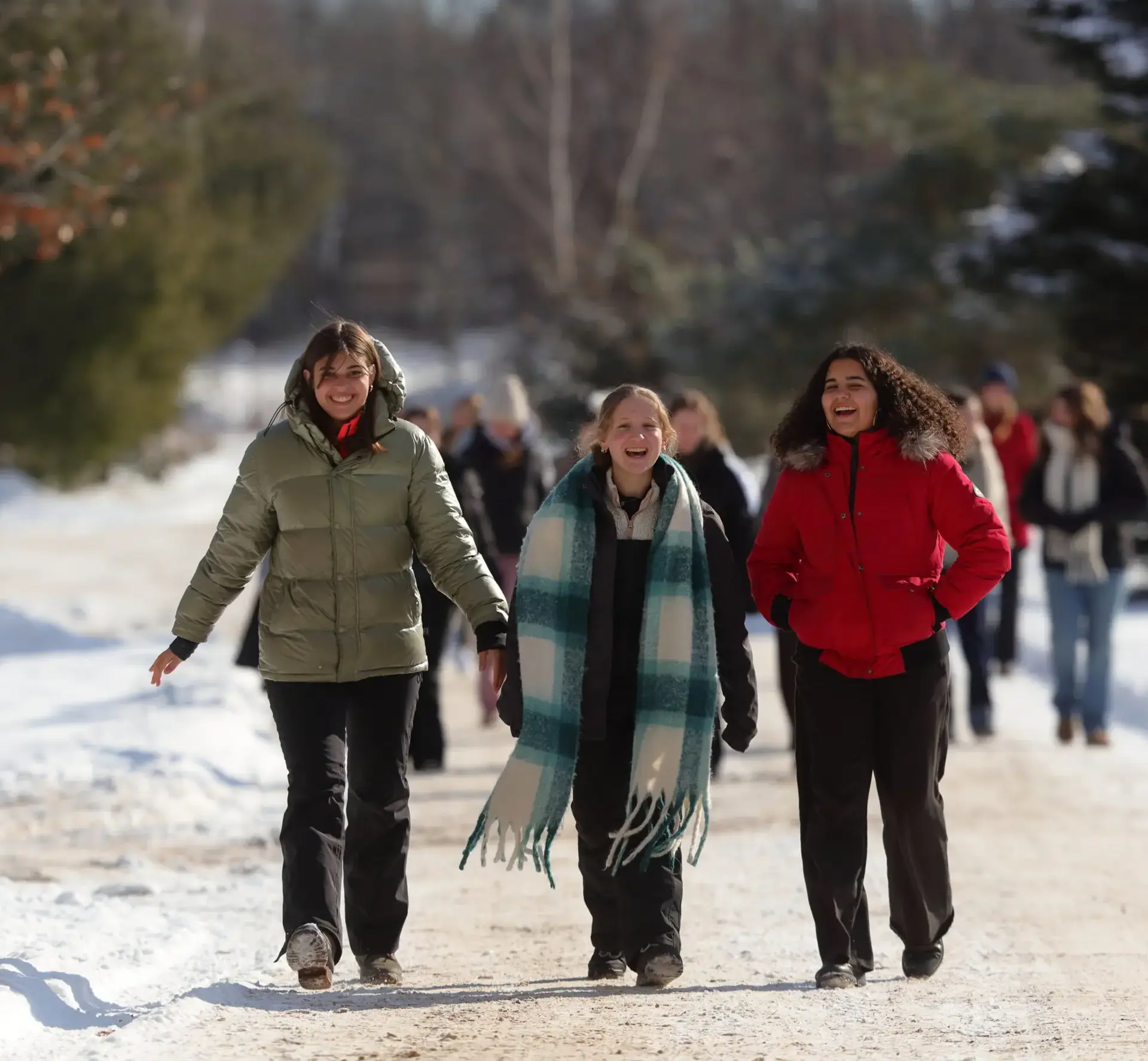 Three students walk side-by-side along a snow-covered path on a bright winter day, laughing and talking.