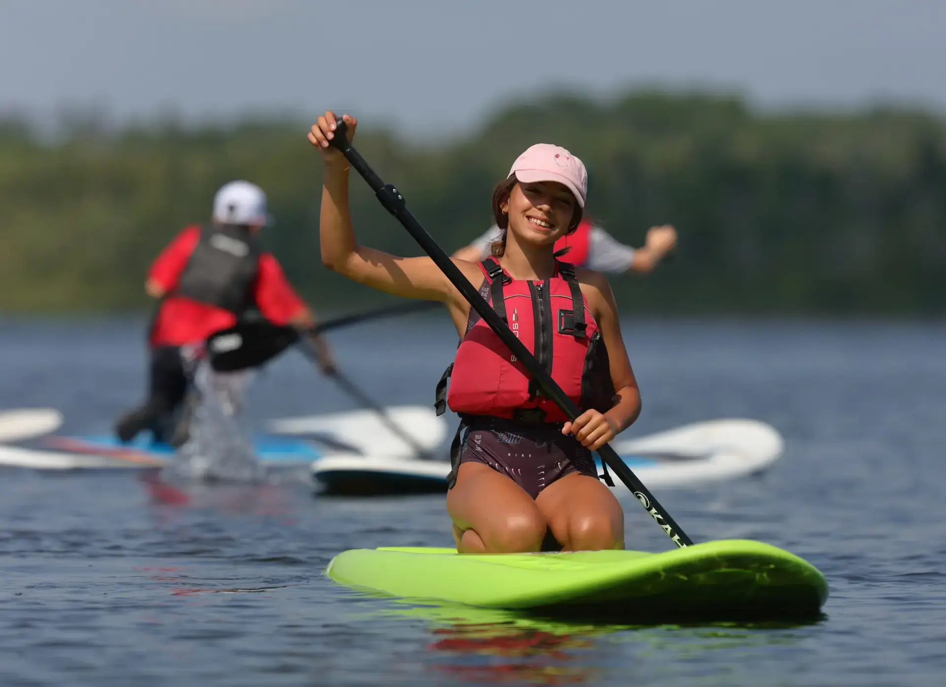 A young girl smiles while kneeling on a bright green paddleboard in the middle of a calm lake.