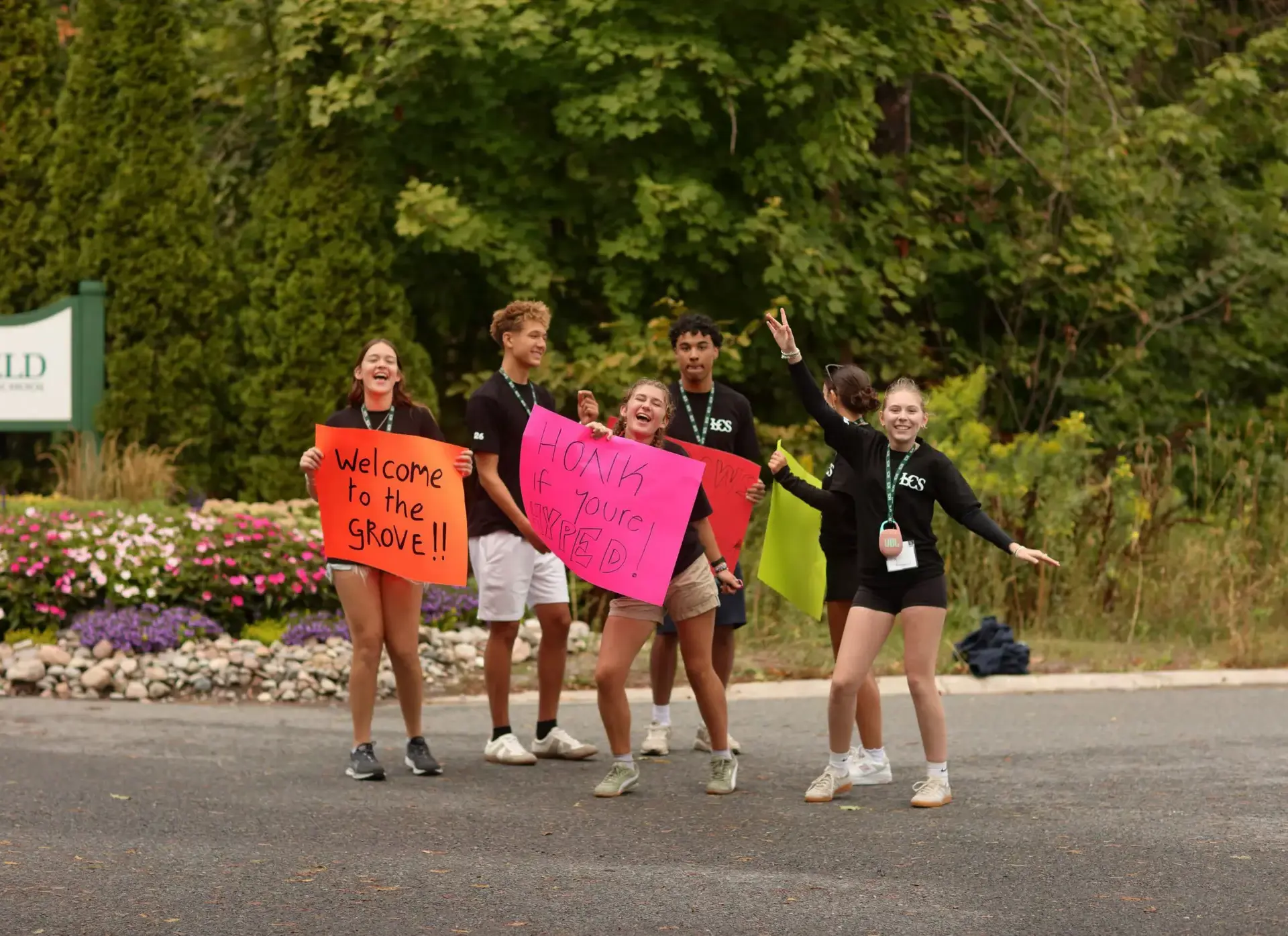 Five students stand together outdoors, holding colorful handmade signs to welcome others to 'the Grove.' 