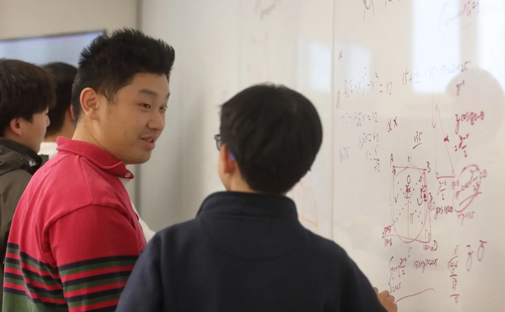 Two students stand at a whiteboard covered in complex mathematical equations and geometric diagrams.