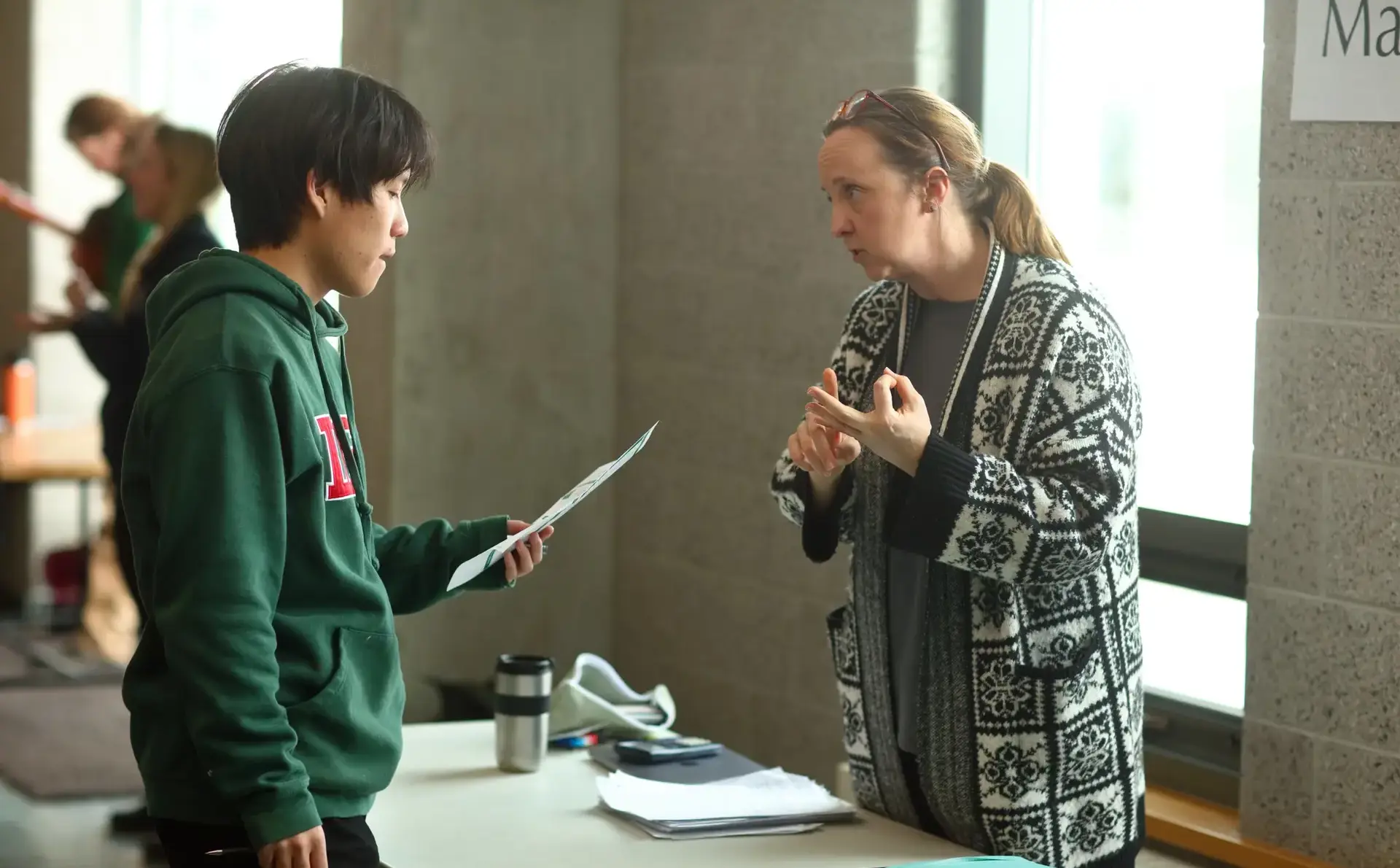 A teacher and a student engage in a focused discussion at a table in a bright hallway.